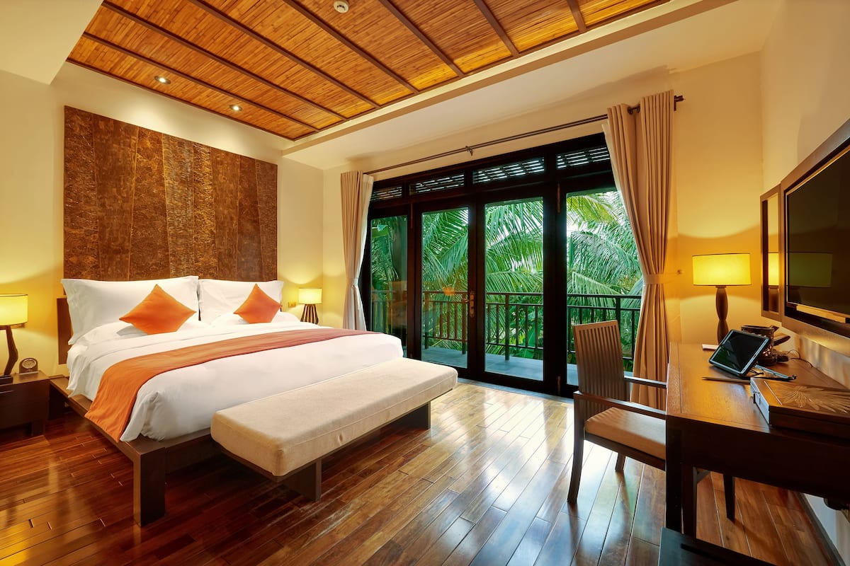 Une chambre d’hôtel moderne dotée d’un grand lit, d’un parquet en bois, d’un bureau avec un ordinateur portable et offrant une vue sur une végétation luxuriante grâce à de grandes portes coulissantes.