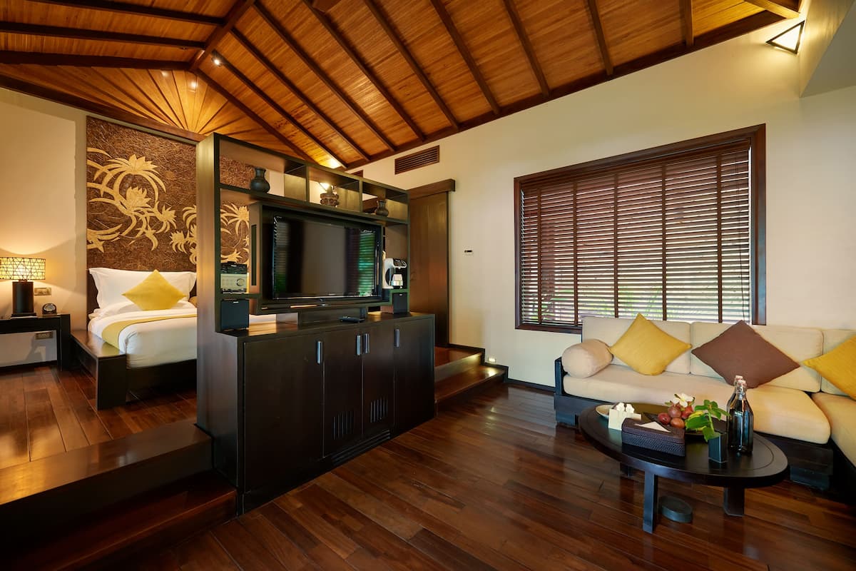 Une chambre d’hôtel moderne avec un plafond en bois, un lit avec des oreillers jaunes, un canapé avec des coussins jaunes et bruns, une table basse avec une bouteille et des fruits, et un téléviseur posé sur un meuble.