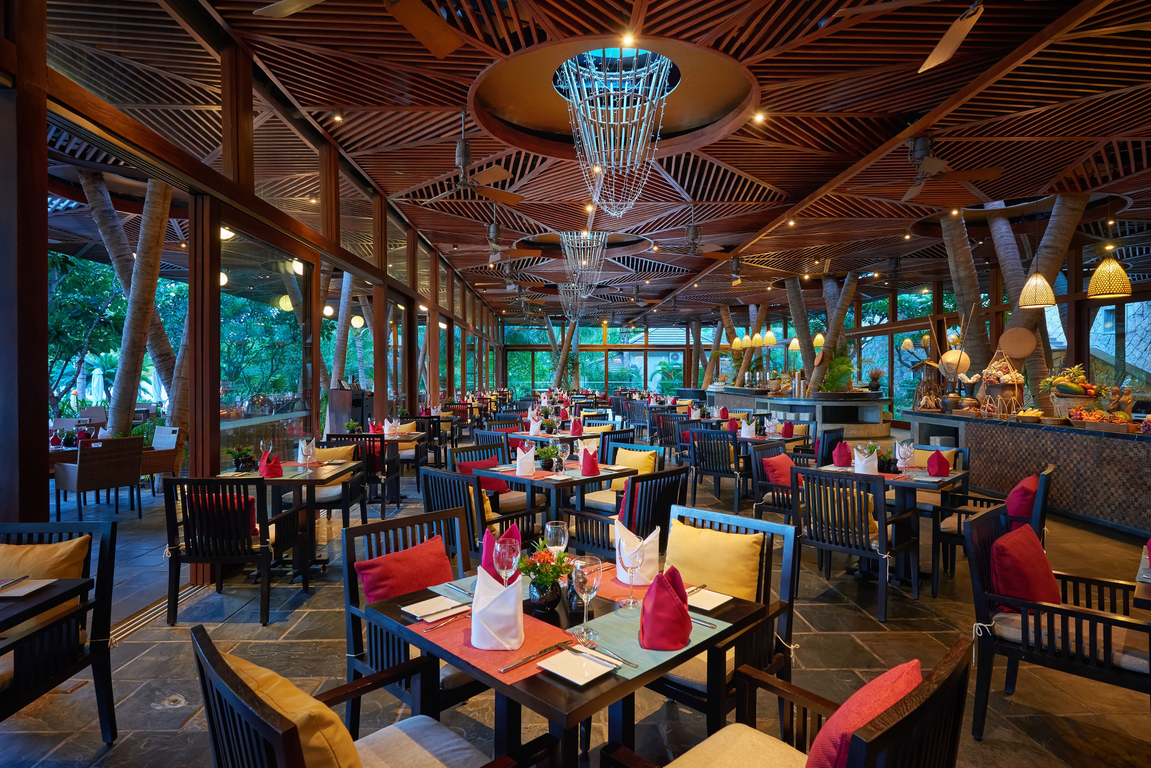 Um restaurante espaçoso com pé-direito alto, vigas de madeira e grandes janelas que oferecem vista para o verde. As mesas estão bem arrumadas com almofadas vermelhas e amarelas, e o interior apresenta um lustre moderno.