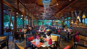 Um restaurante espaçoso com pé-direito alto, vigas de madeira e grandes janelas que oferecem vista para o verde. As mesas estão bem arrumadas com almofadas vermelhas e amarelas, e o interior apresenta um lustre moderno.