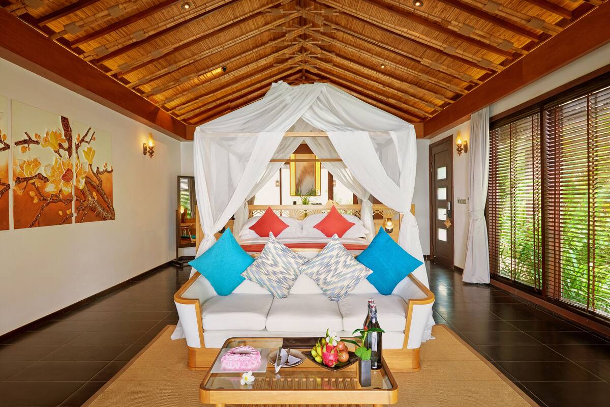 Une chambre avec un lit à baldaquin, un canapé blanc et une table basse sur laquelle se trouvent des fruits et une bouteille.