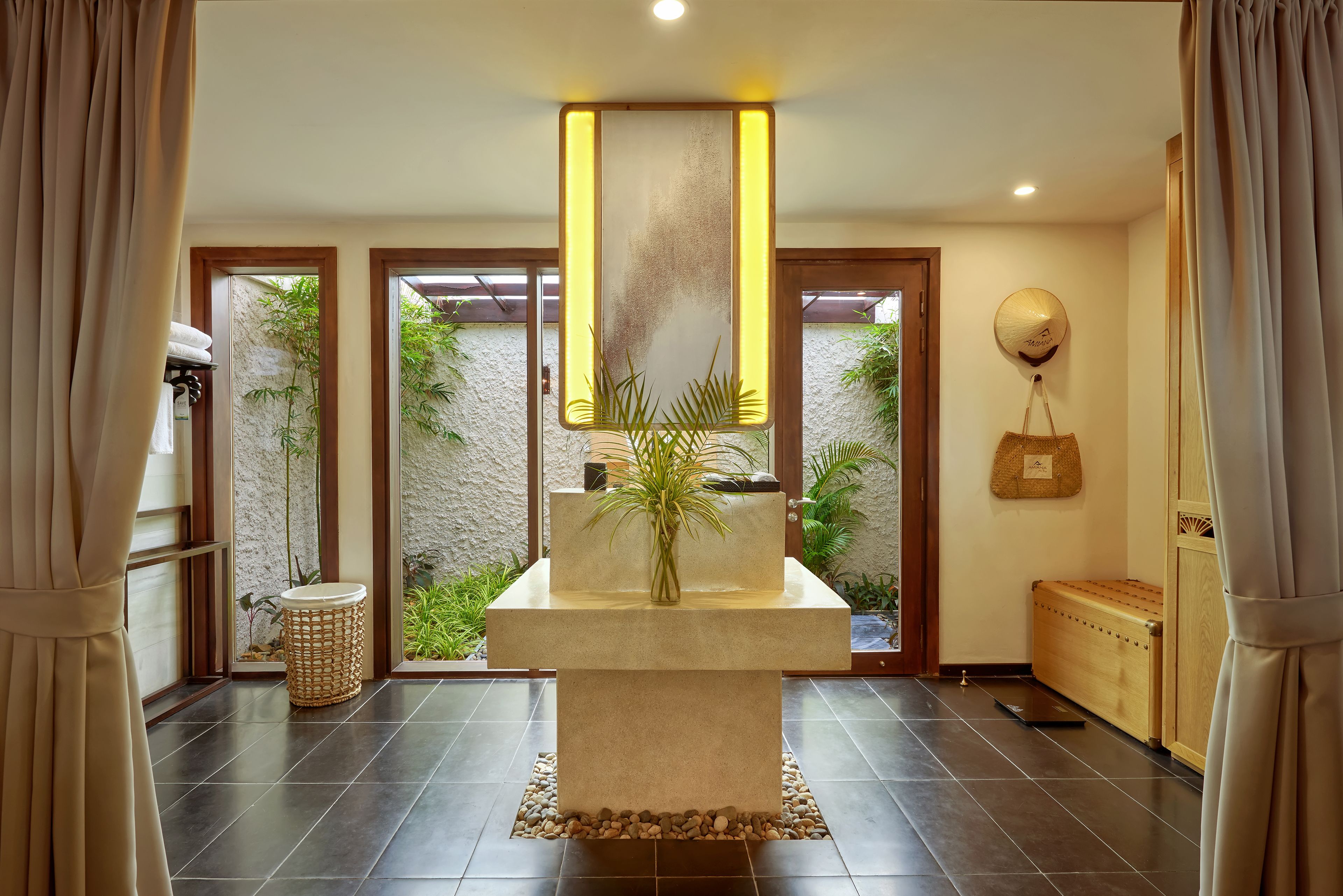 A modern interior with a central stone table, a hanging light fixture, and a view of greenery outside.