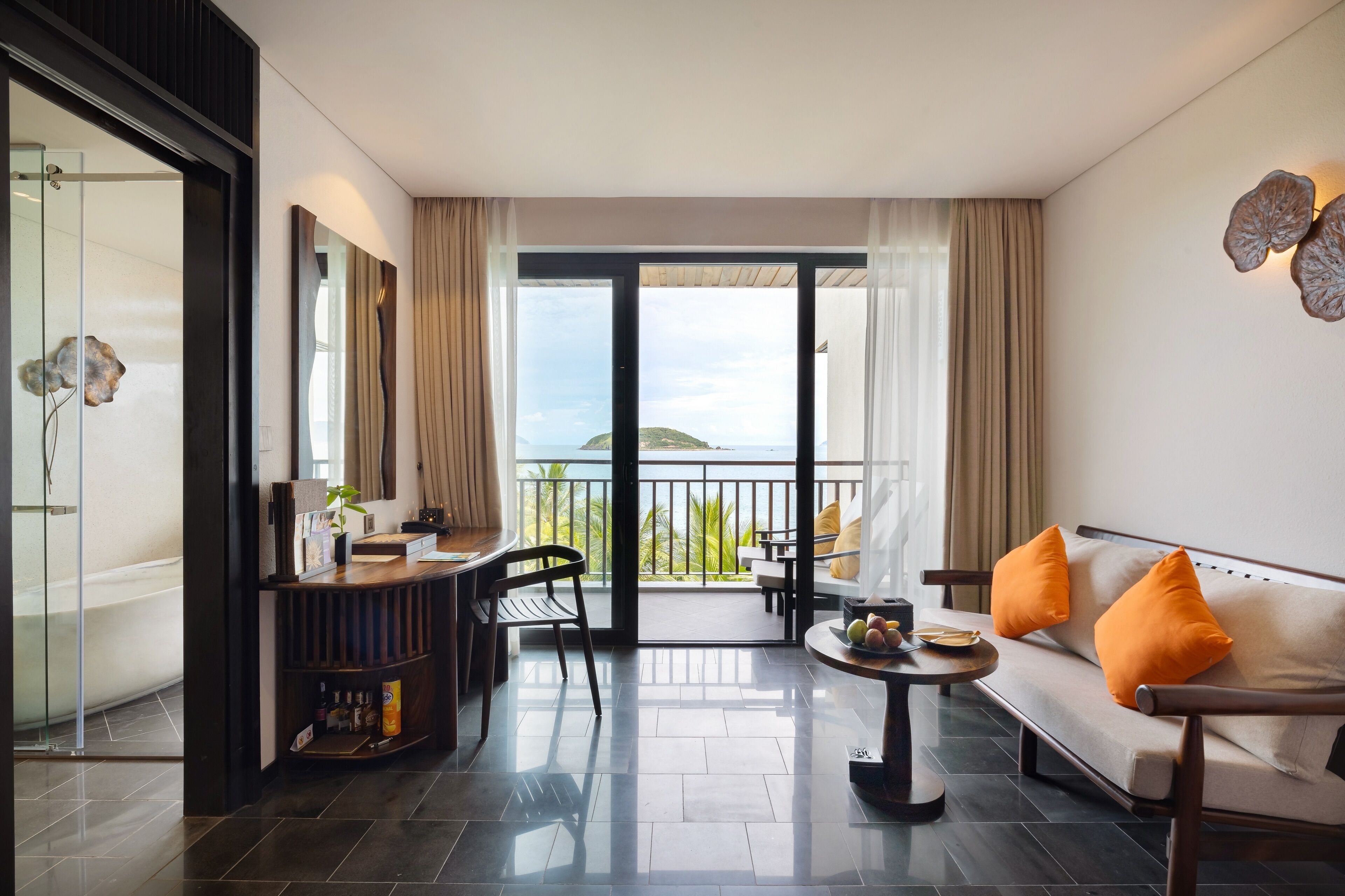 A modern hotel room with a balcony, a sofa with orange cushions, a dining area with a table and chairs, and a large mirror.