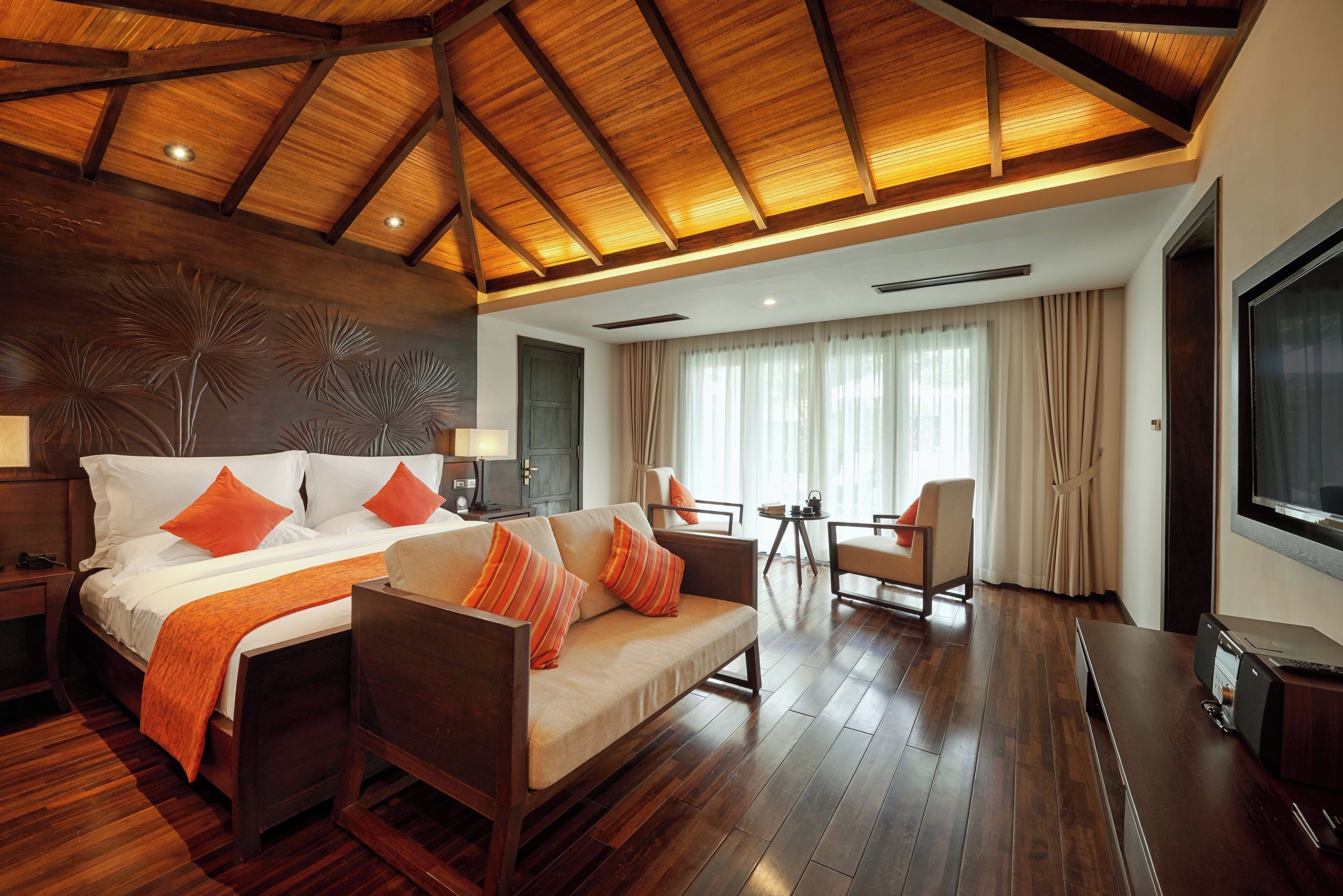 Una habitación amplia con techo de madera, una cama grande con almohadas naranjas, un sofá, un televisor y una zona de comedor con mesa y sillas.