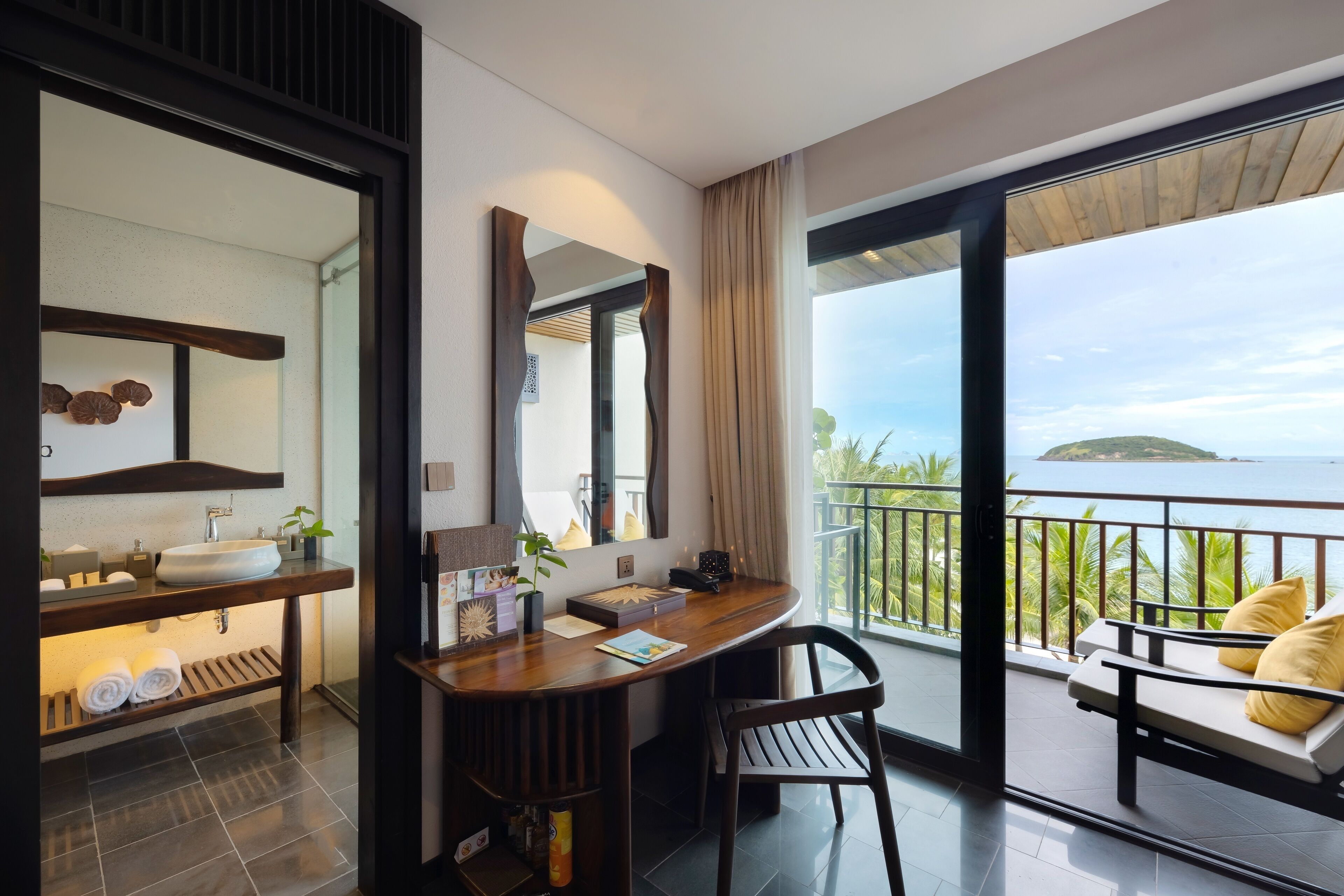 Une salle de bain moderne avec un grand miroir, un meuble-lavabo en bois et un évier. On y trouve également un bureau avec une chaise et une vue sur la mer depuis un balcon.