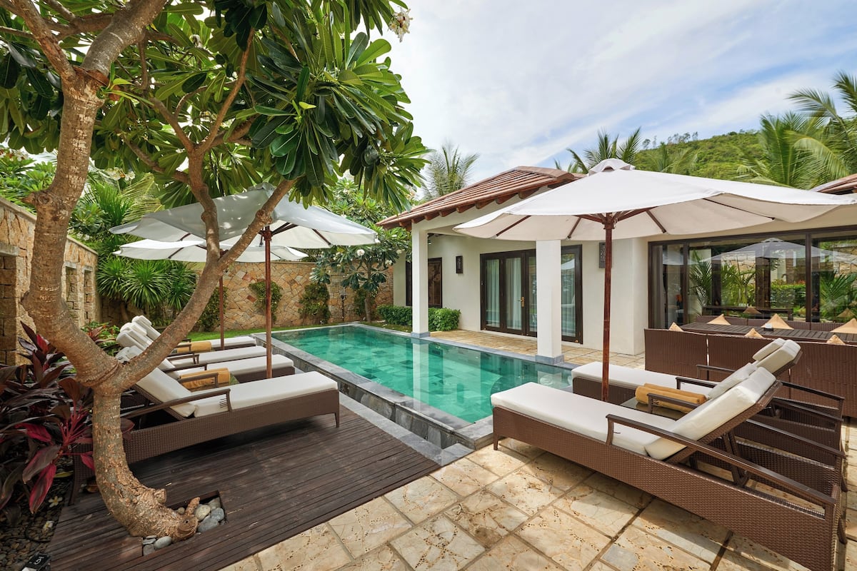 Un espace aménagé au bord de la piscine, avec des chaises longues et des parasols, entouré de verdure et délimité par un mur en pierre.