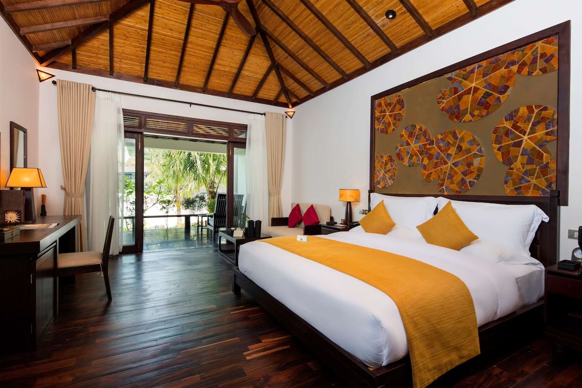 Une chambre d’hôtel avec un grand lit, un plancher en bois, un bureau avec une lampe et une vue sur l’extérieur, visible à travers une porte vitrée.