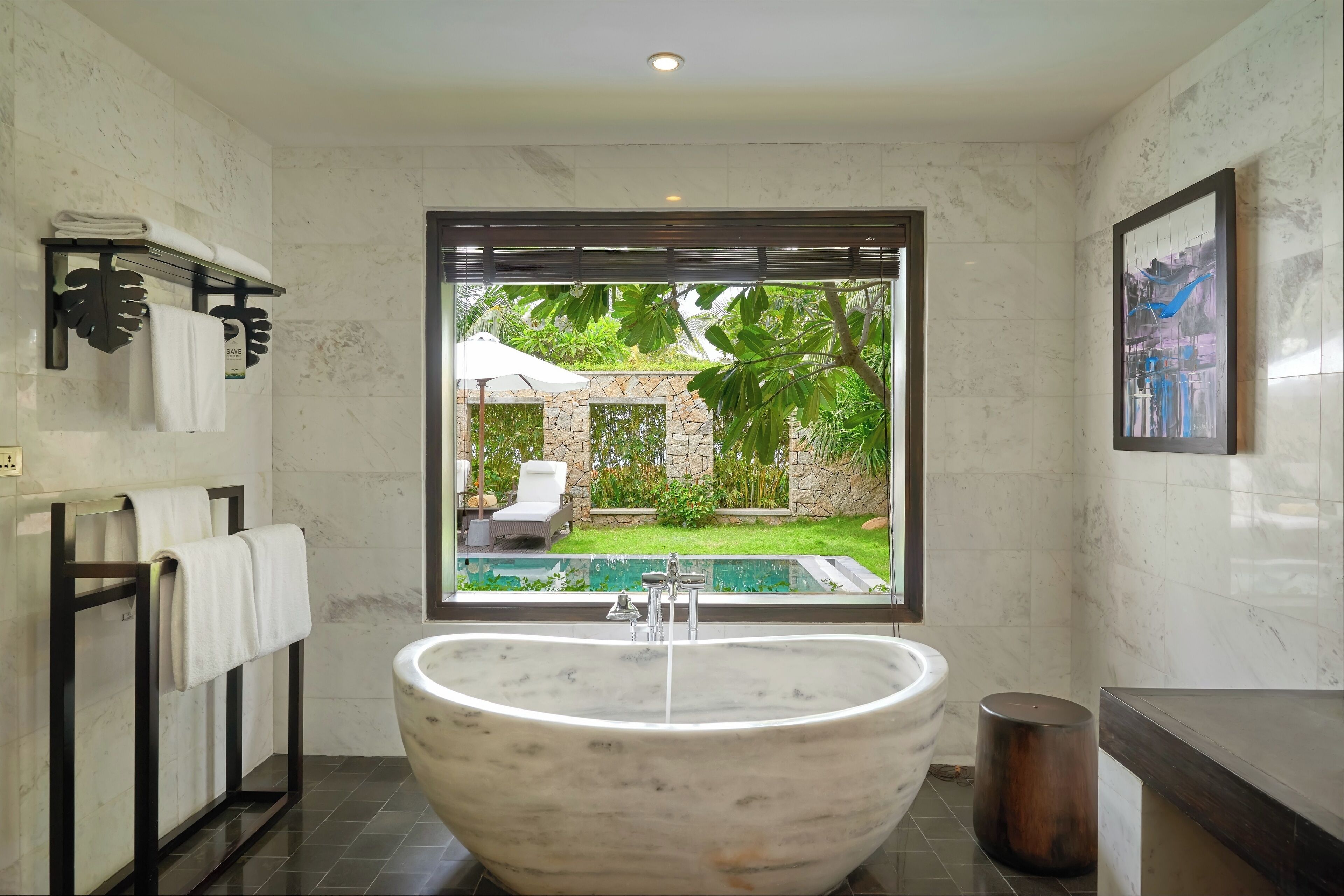 A modern bathroom with a large freestanding bathtub, a window with a view of a pool and greenery, and a framed artwork on the wall.