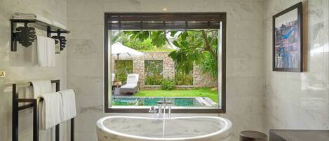 A modern bathroom with a large freestanding bathtub, a window with a view of a pool and greenery, and a framed artwork on the wall.