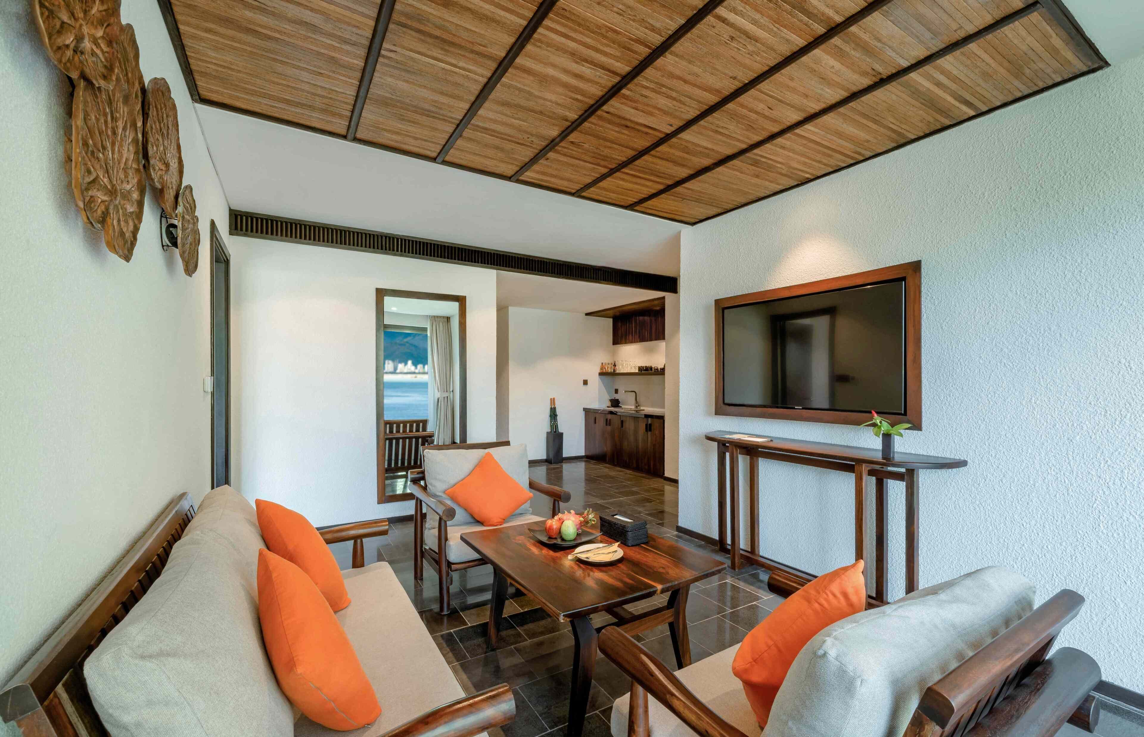 A living area with a wooden ceiling, a TV mounted on the wall, a dining table with chairs, and a view of the sea through a window.