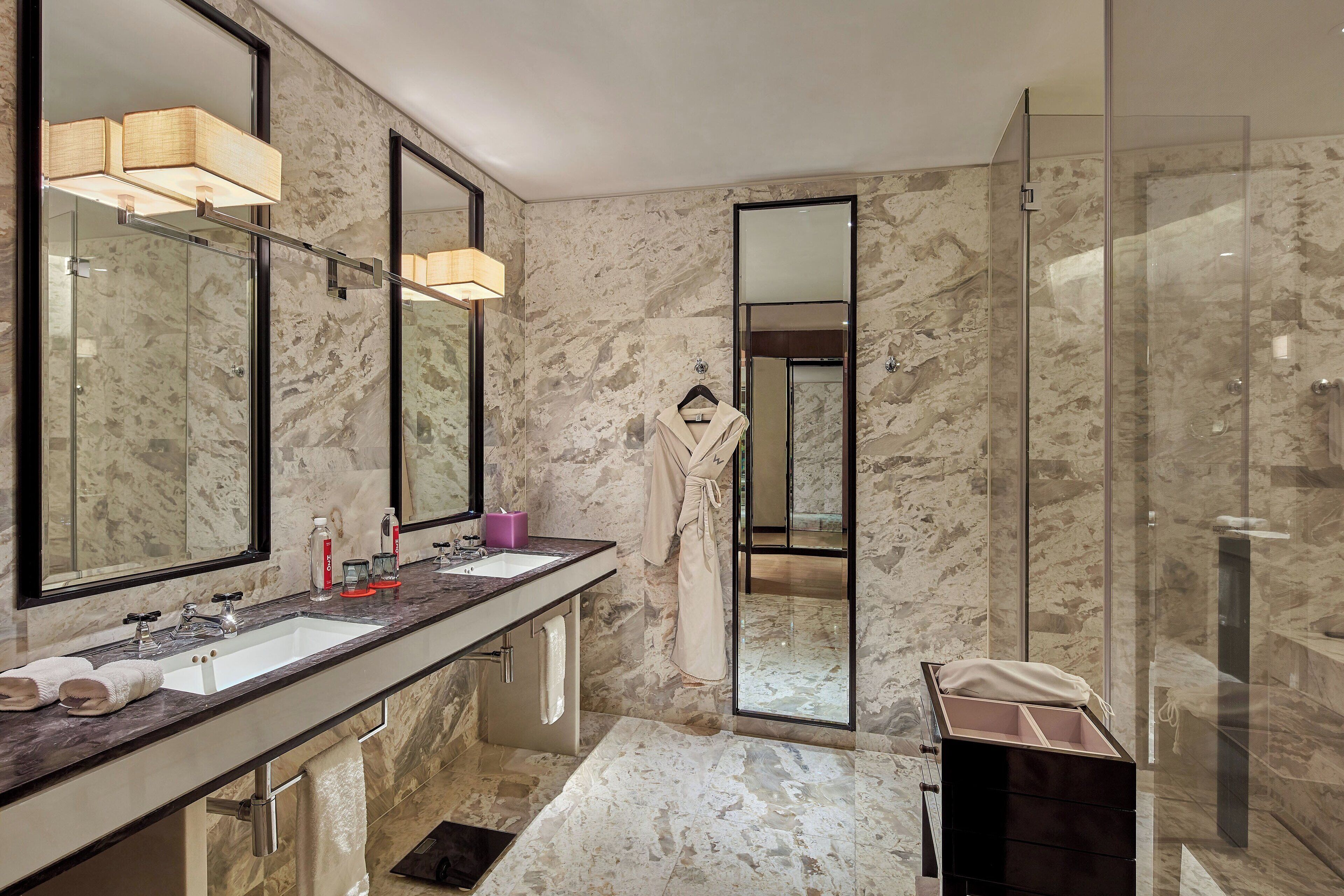 Un baño moderno con paredes de mármol, un espejo grande, cabina de ducha de vidrio y tocador con lavamanos y toallero.