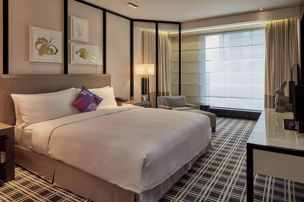 Une chambre d’hôtel avec un grand lit, un bureau avec une chaise, une table de chevet, une lampe et une grande fenêtre avec des rideaux.