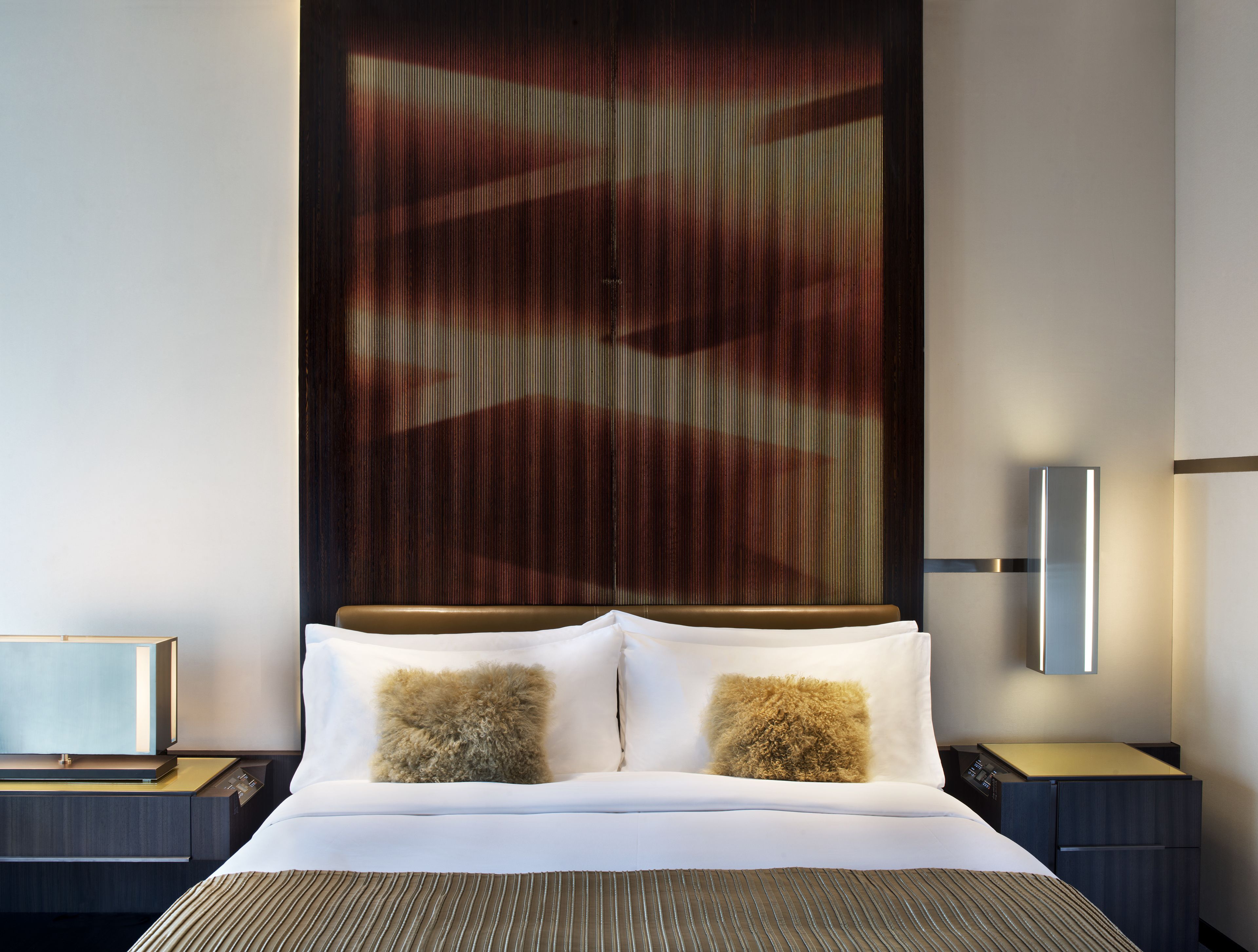 A modern hotel room with a large bed, two pillows, a bedside table, and a wall-mounted lamp.