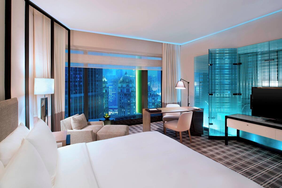 Une chambre d’hôtel dotée d’un grand lit, d’un bureau, d’une chaise, d’une télévision et offrant une vue sur le paysage urbain grâce à de grandes fenêtres du sol au plafond.