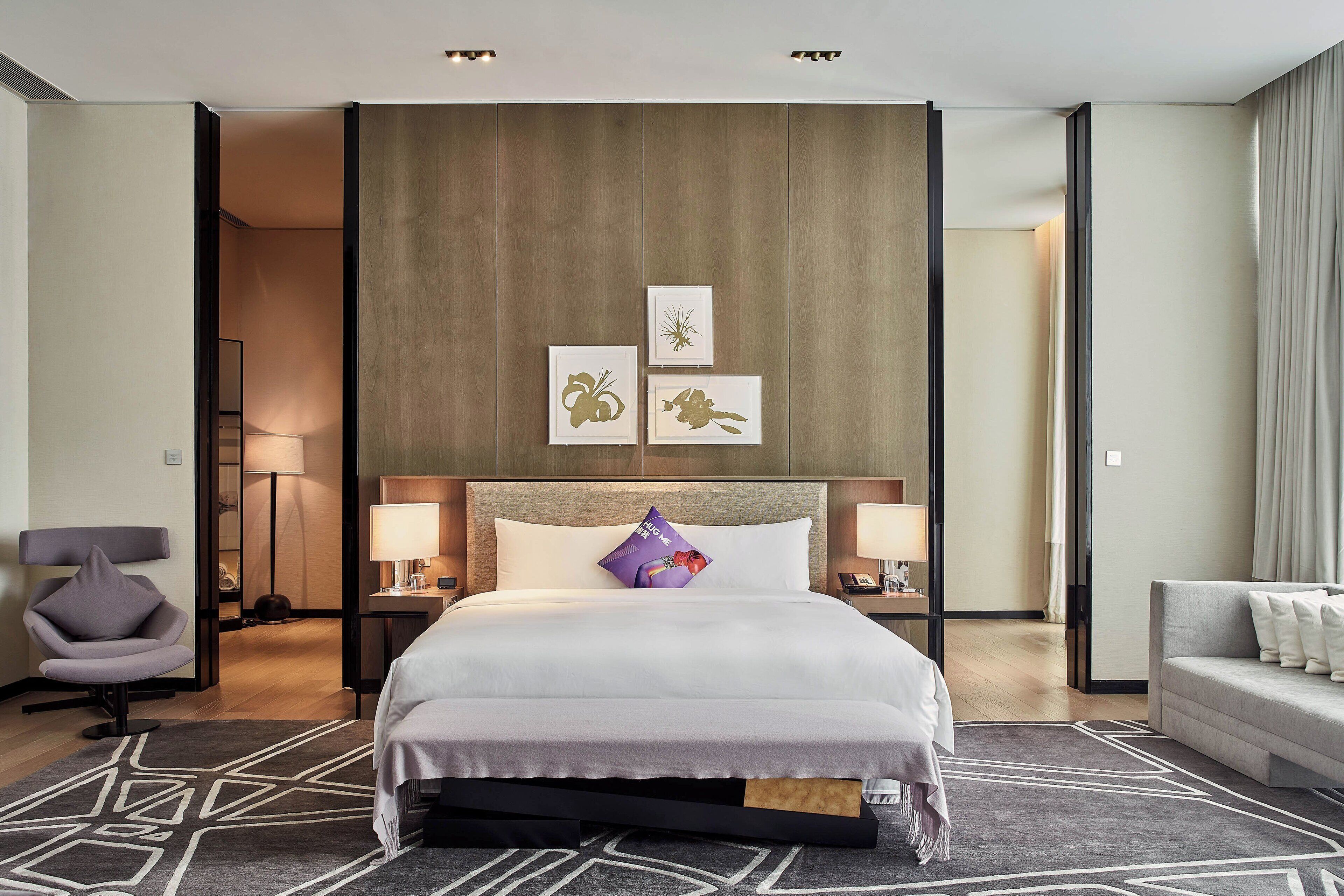 Una habitación de hotel moderna con una cama grande, dos mesitas de noche, una silla, un sofá y obras de arte en la pared.
