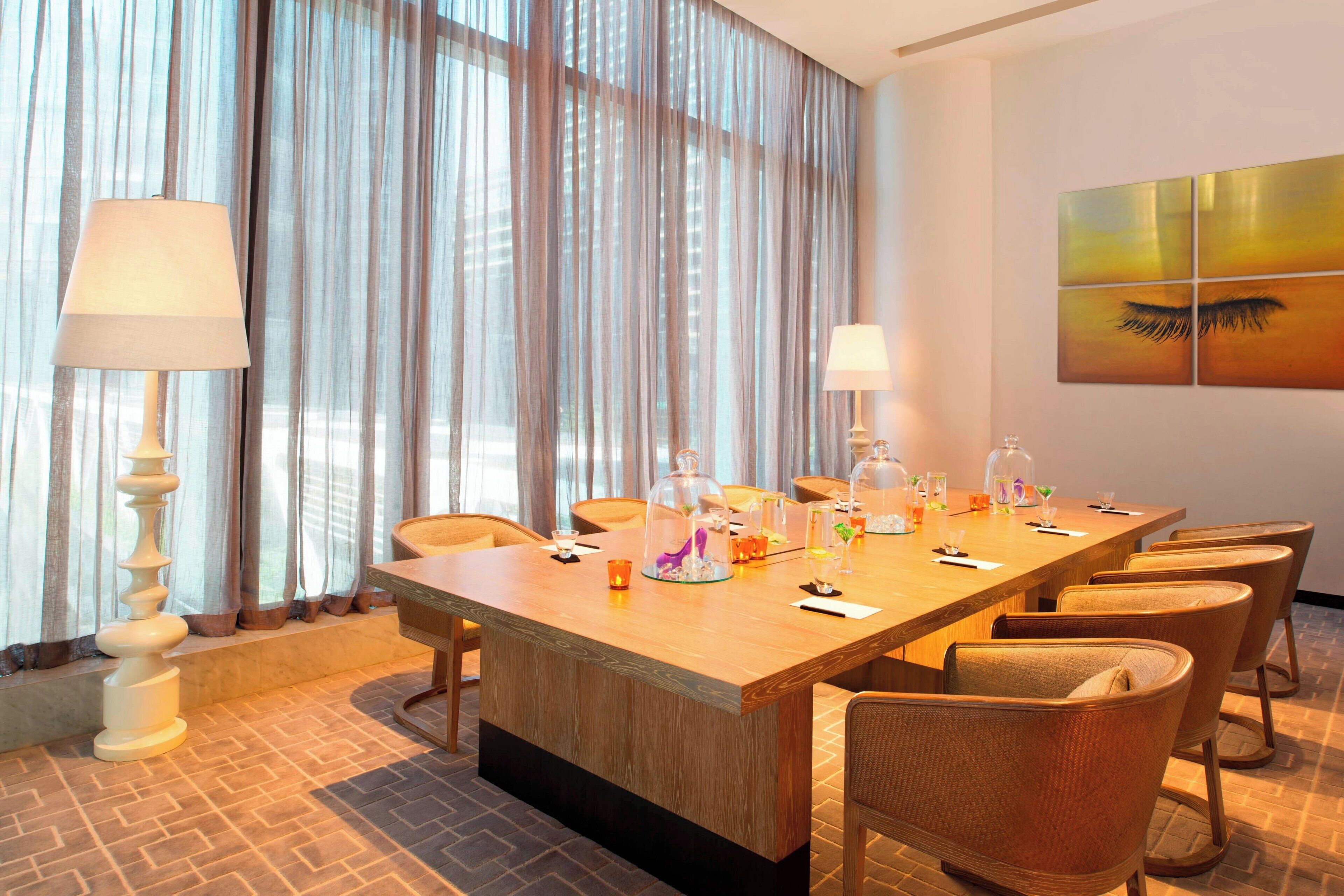 Een hotelconferentiezaal met een lange houten tafel, zes stoelen en een groot raam met dunne gordijnen.