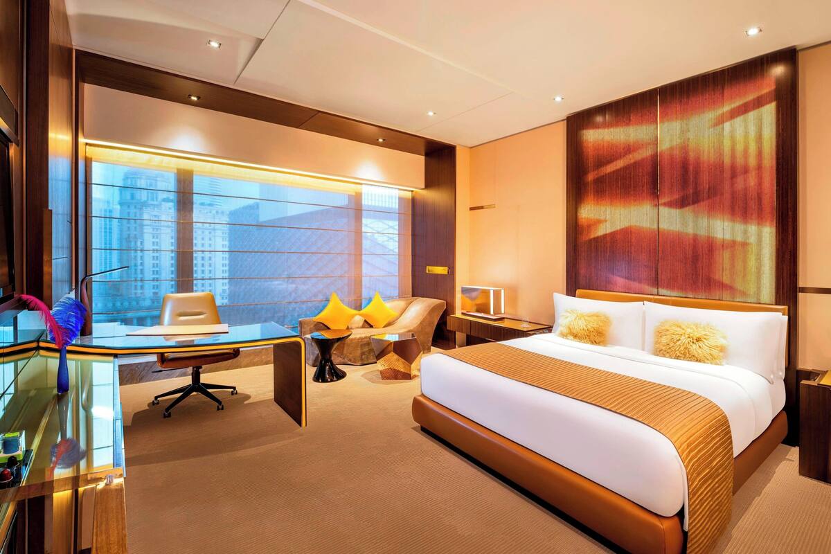 Une chambre d’hôtel moderne avec un grand lit, un bureau et une chaise. De grandes fenêtres offrent une vue sur la ville.