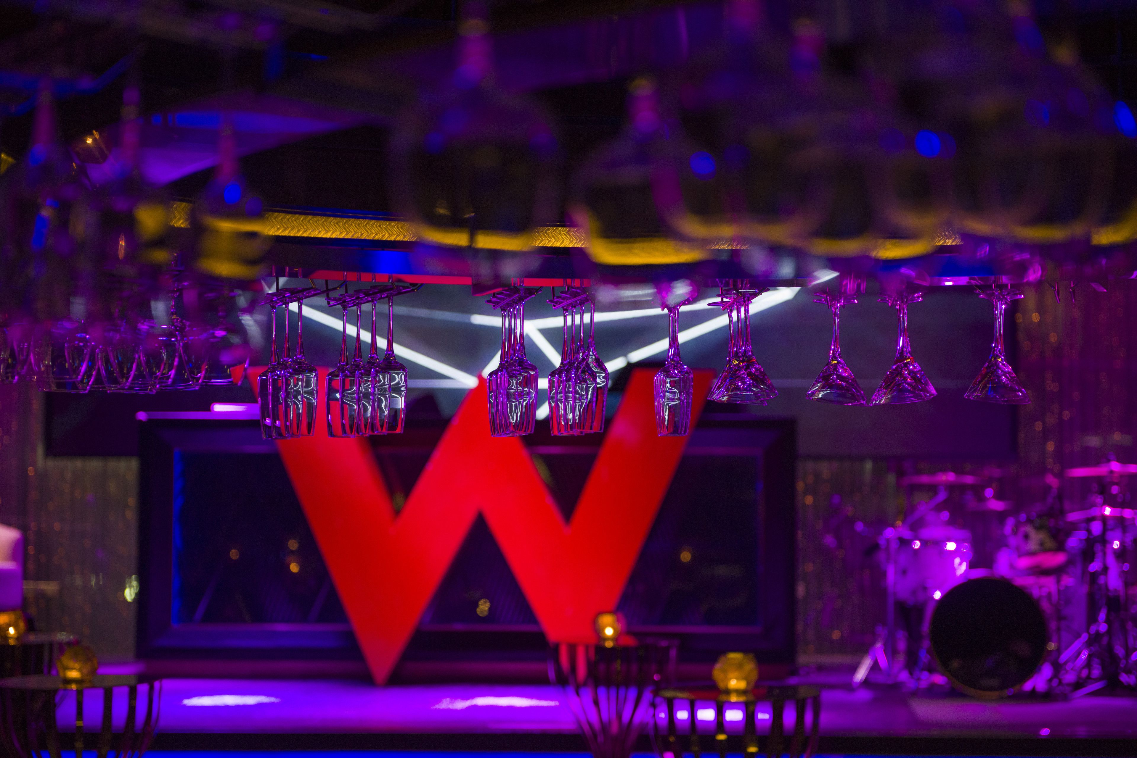 A stage with a large illuminated 'W' sign, multiple hanging crystal glasses, and a drum set in the background.
