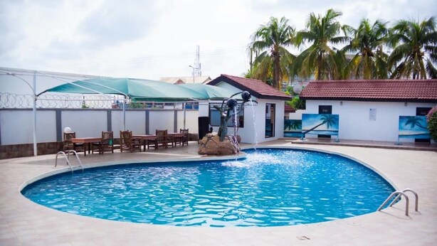 A swimming pool with a water fountain, surrounded by palm trees and a covered seating area. - Rexmar Hotel (Kumasi)