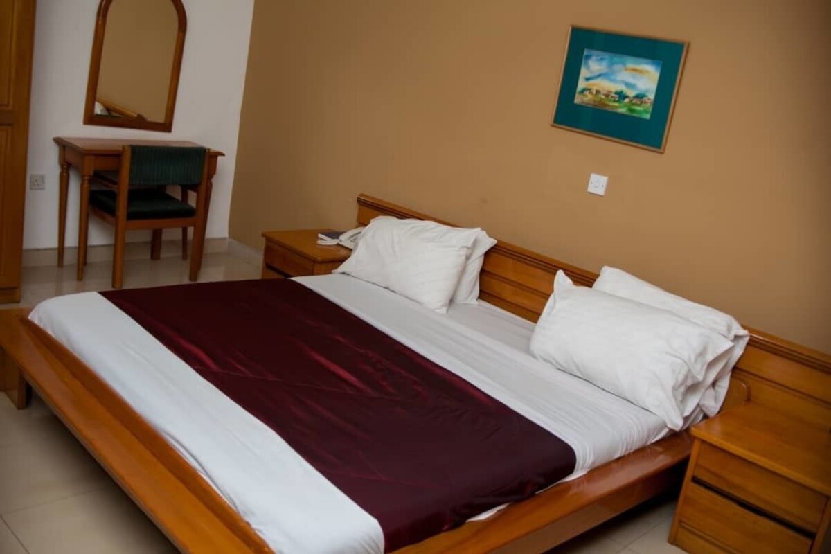 A neatly made bed with a wooden frame and white bedding, a wooden nightstand with a lamp, a chair with a green cushion, and a painting on the wall.