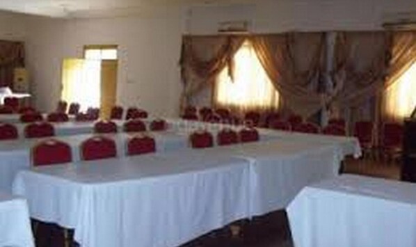 Un salón de conferencias con varias mesas y sillas, amplias ventanas con cortinas y una puerta visible.