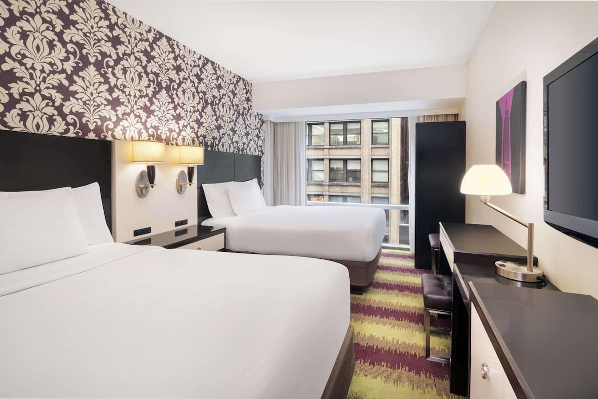 A hotel room with two beds, a desk, a chair, a TV, and a window with a city view.