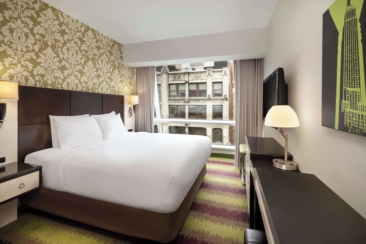 A hotel room with a large bed, a desk, a TV, and a window with a city view.