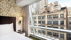 A hotel room with a large bed, a nightstand with a lamp, and a view of a cityscape through a large window. - Best Western Premier Herald Square (New York)