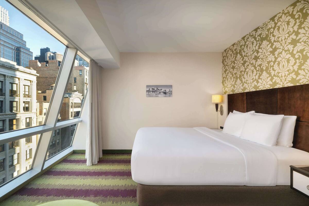 A hotel room with a large bed, a view of the city through a window, a patterned headboard, and a framed picture on the wall.