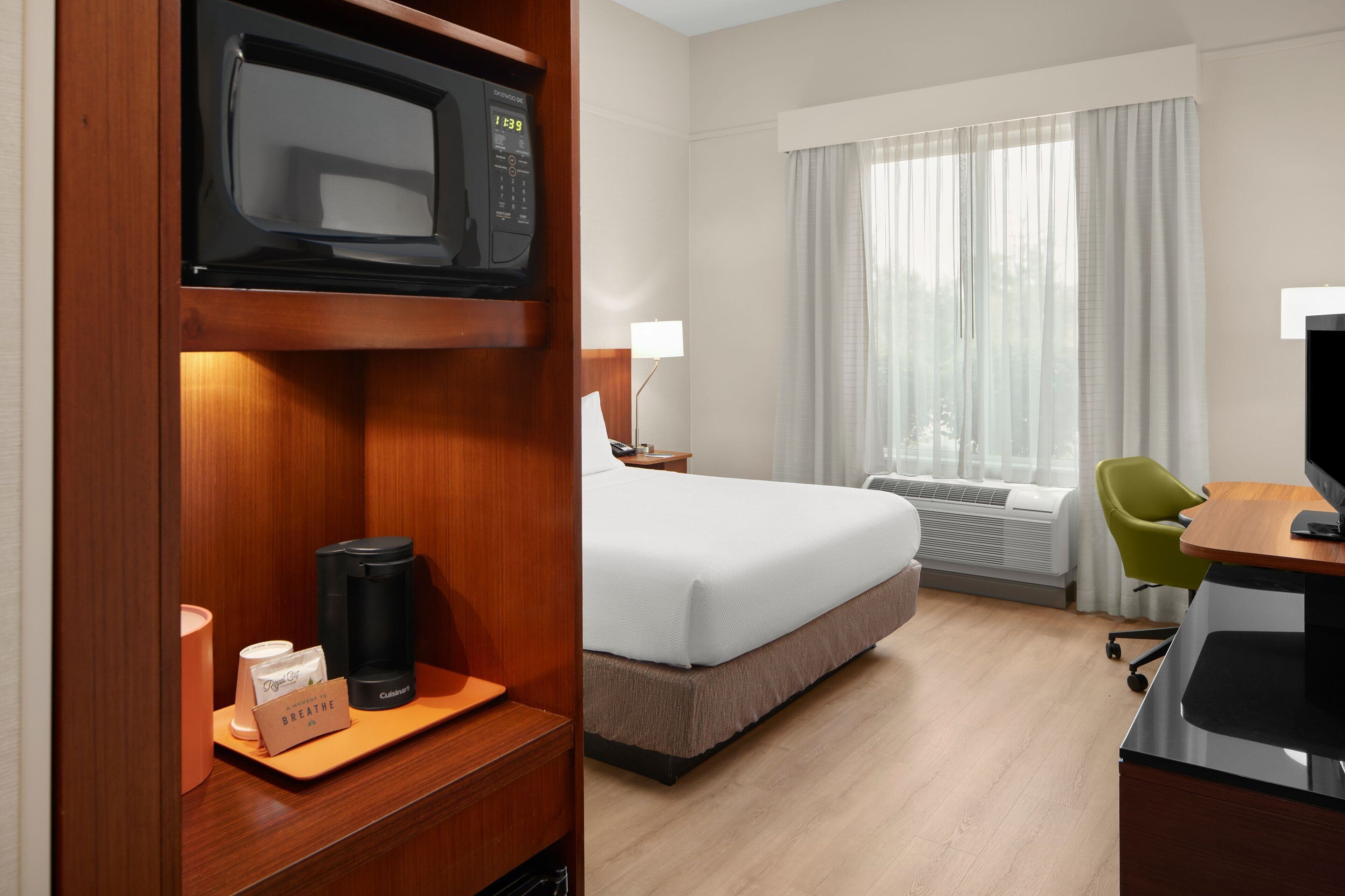 A hotel room with a bed, a desk with a computer, a microwave, a coffee maker, and a window with curtains.