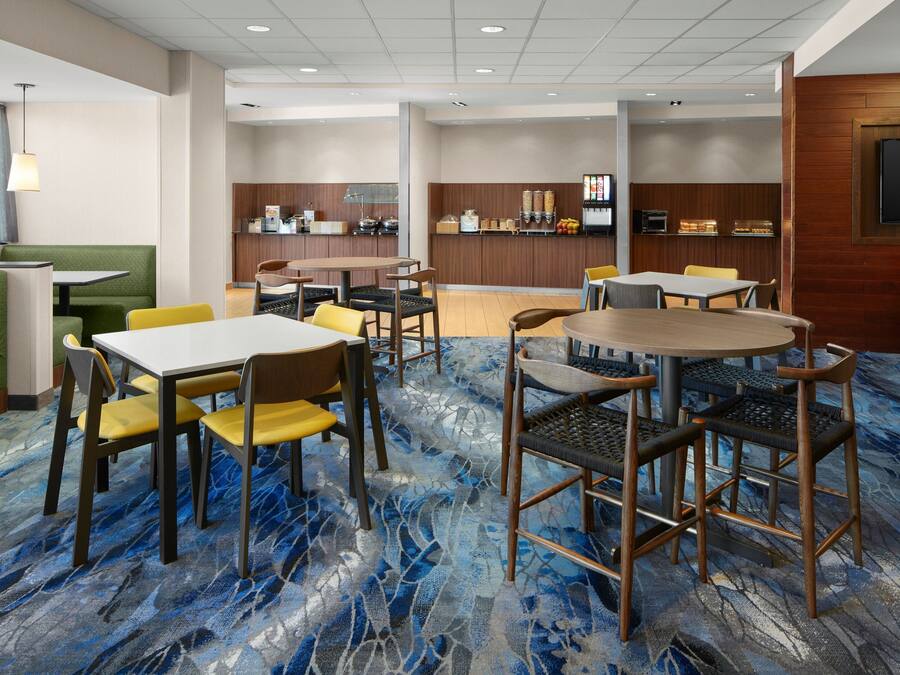 Een hotel lobby met diverse zitmogelijkheden, waaronder zitjes, tafels en stoelen. Er is een receptiebalie met een aanrecht en een televisie aan de muur. De vloer heeft een blauw-wit betegeld tapijt.
