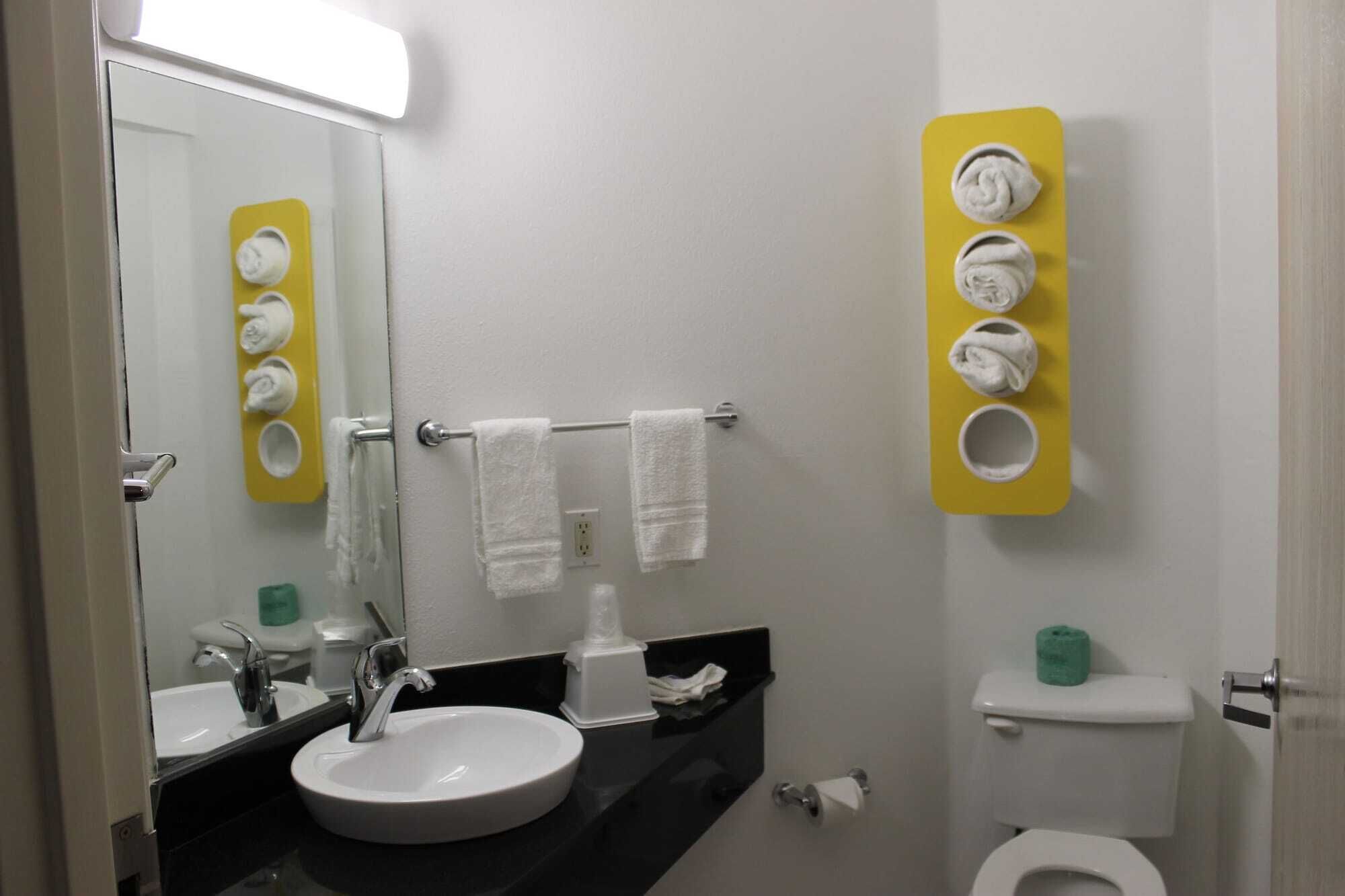 A bathroom with a white sink, mirror, and towel rack. There is a toilet with a green toilet paper holder and a yellow towel dispenser.