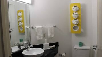 Un baño con lavamanos blanco, espejo y perchero para toallas. Hay un inodoro con un dispensador verde de papel higiénico y un dispensador amarillo de toallas.