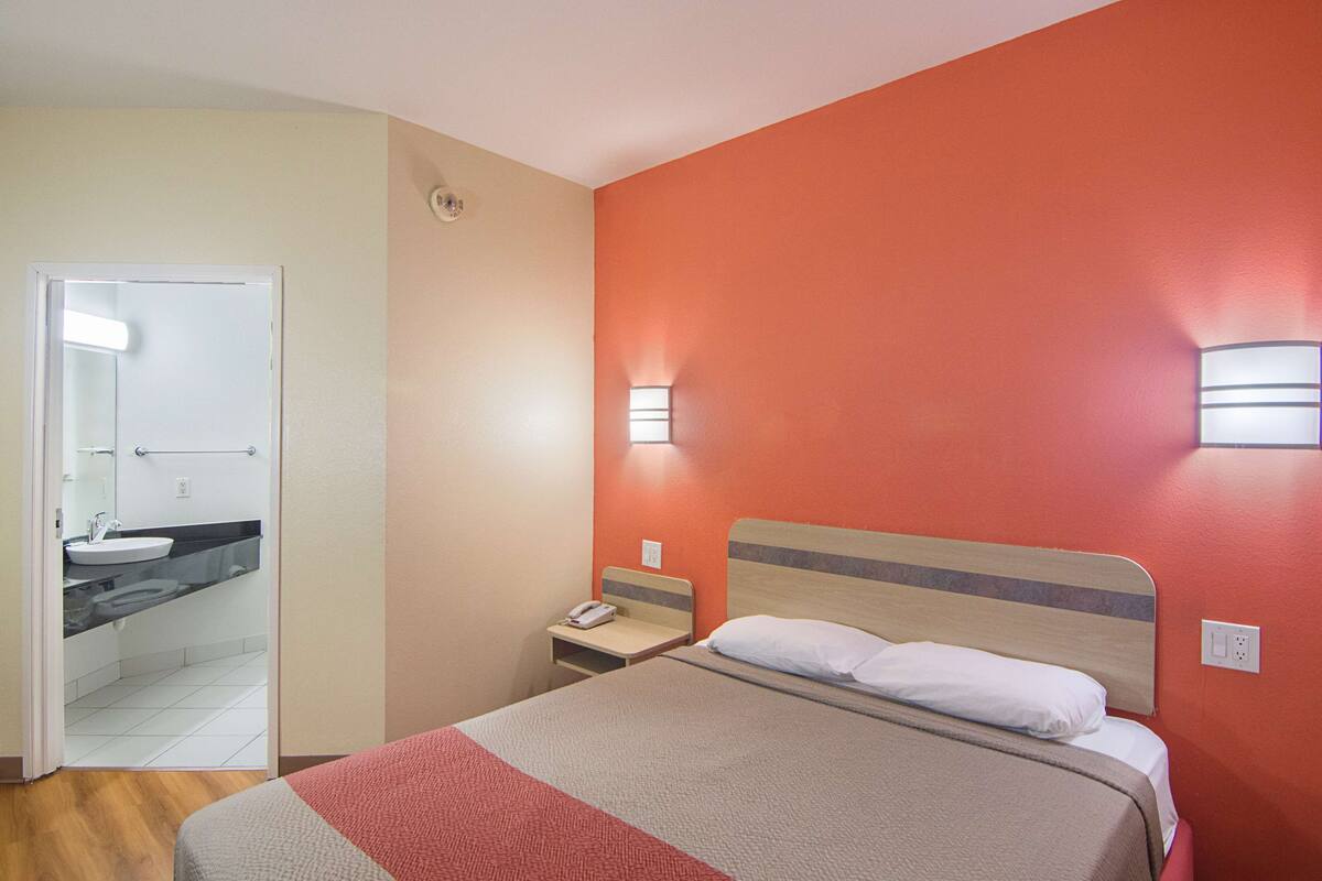 Habitación de hotel con cama, mesita de noche, teléfono y baño visible a través de una puerta abierta.