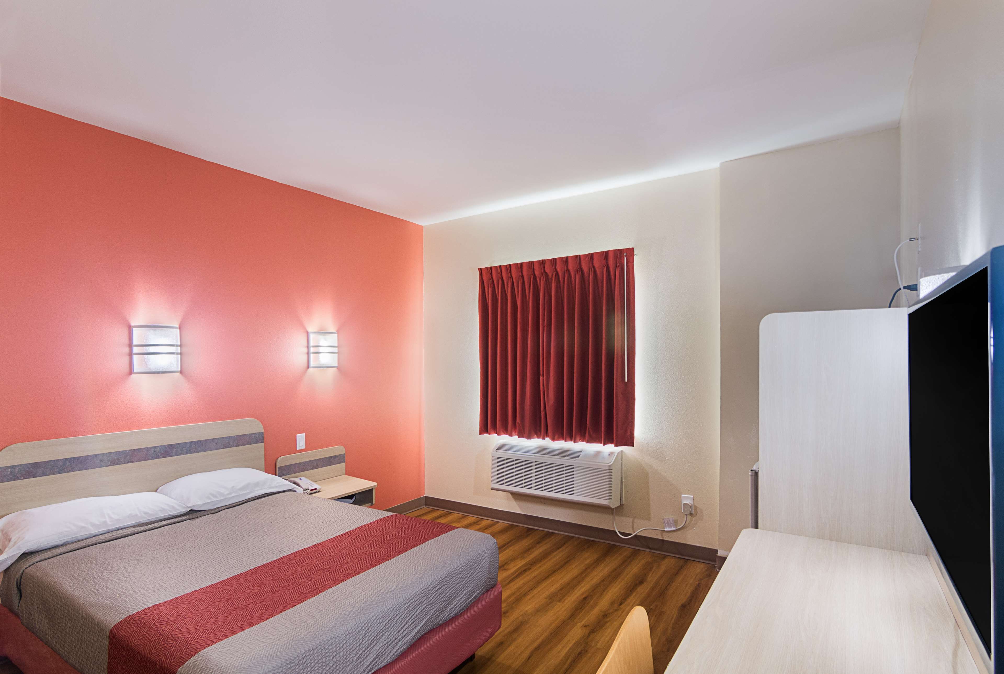 A hotel room with a bed, bedside table, lamp, window with curtains, and a TV.