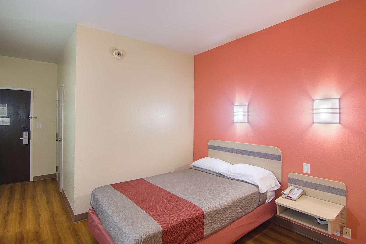Una cama individual con cabecera de madera, una mesita de noche con un teléfono y una lámpara de pared.