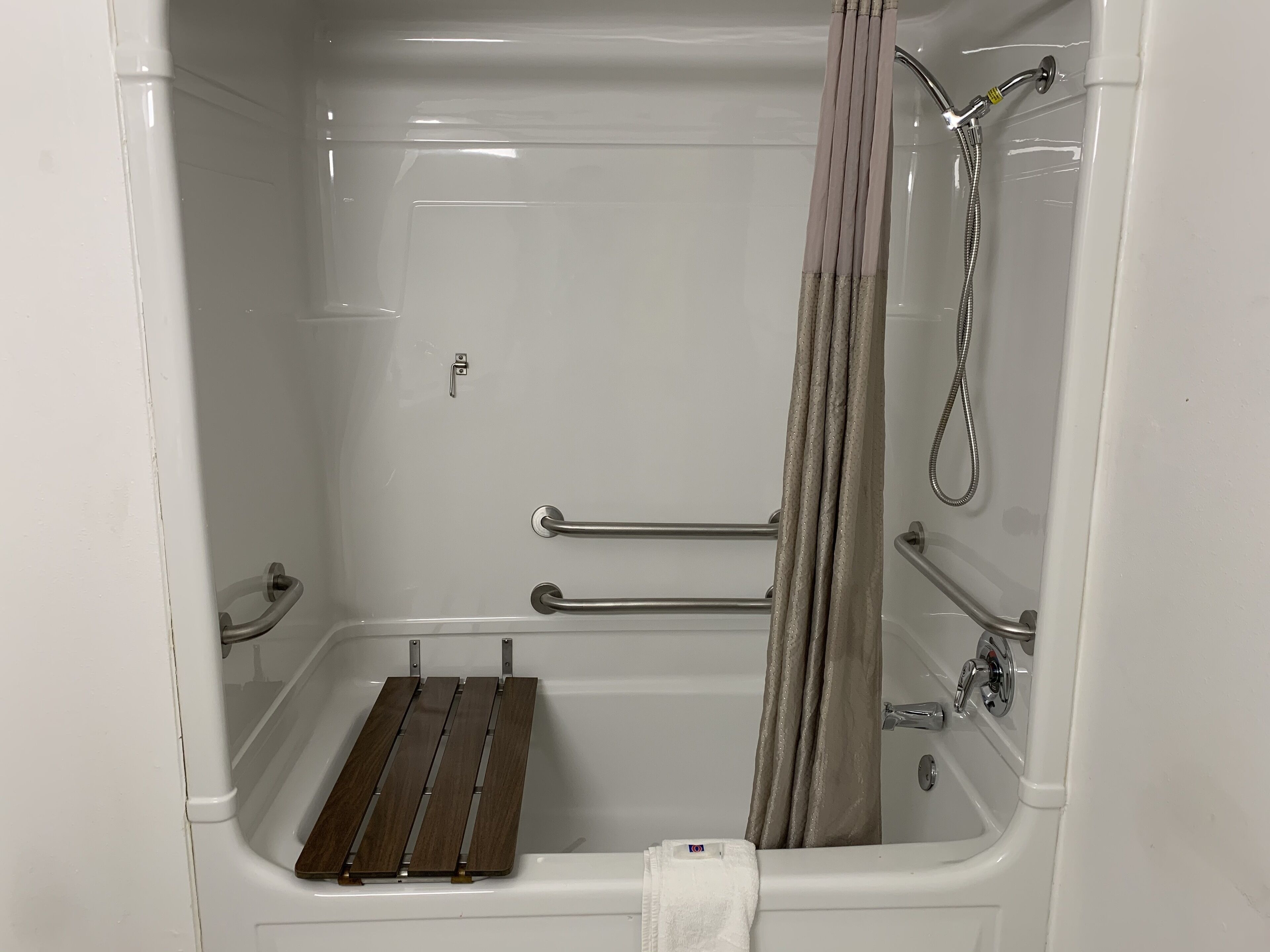 Eine weiße Badewanne mit Holzbank, Haltegriffen und Duschkopf.