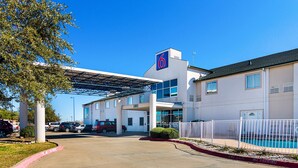A motel with a covered parking area, a swimming pool, and a clear blue sky. - Motel 6 Terrell, TX (Terrell)