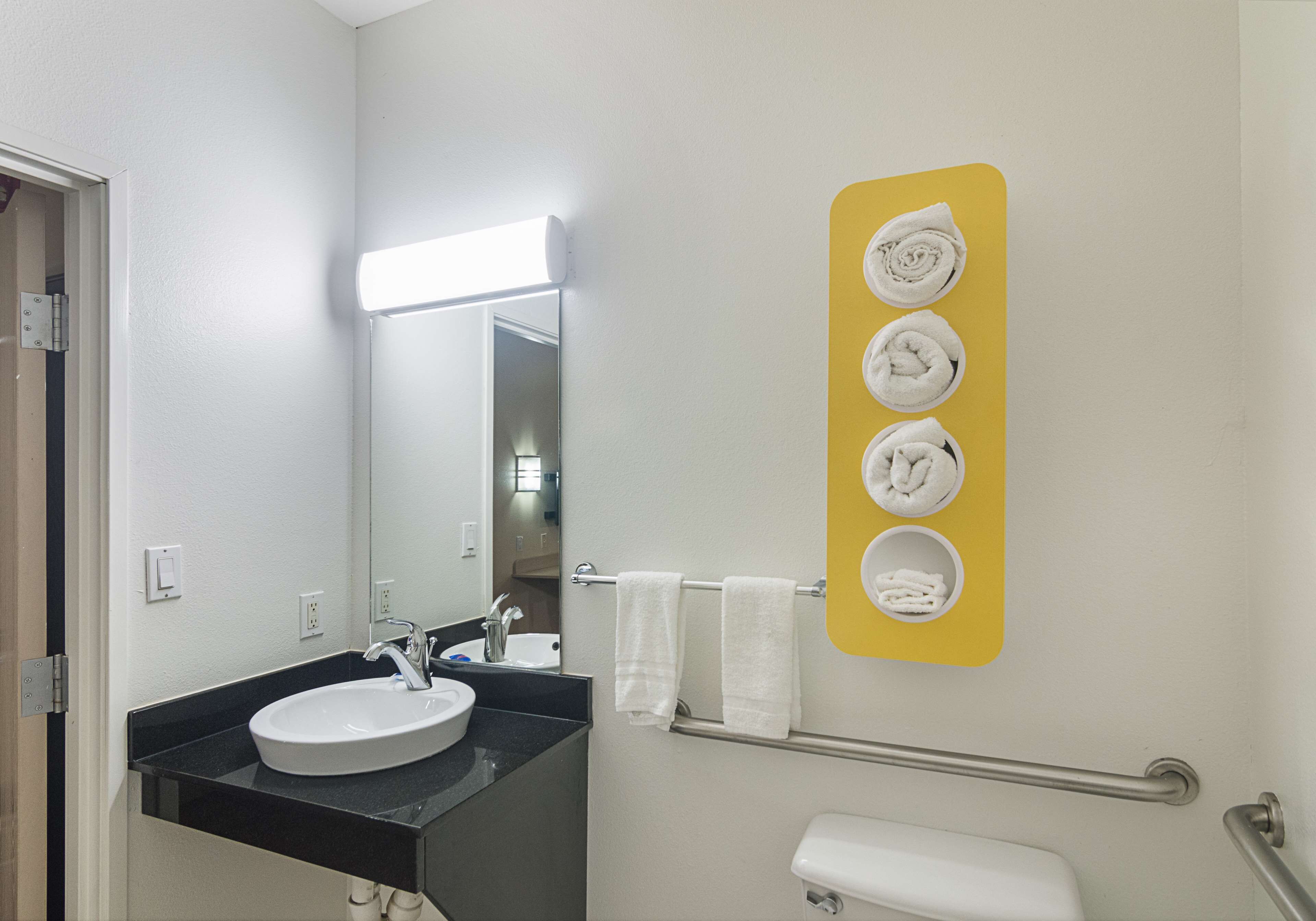 A bathroom with a white sink, a mirror, a towel rack with two towels, a toilet, and a yellow sign with images of towels.