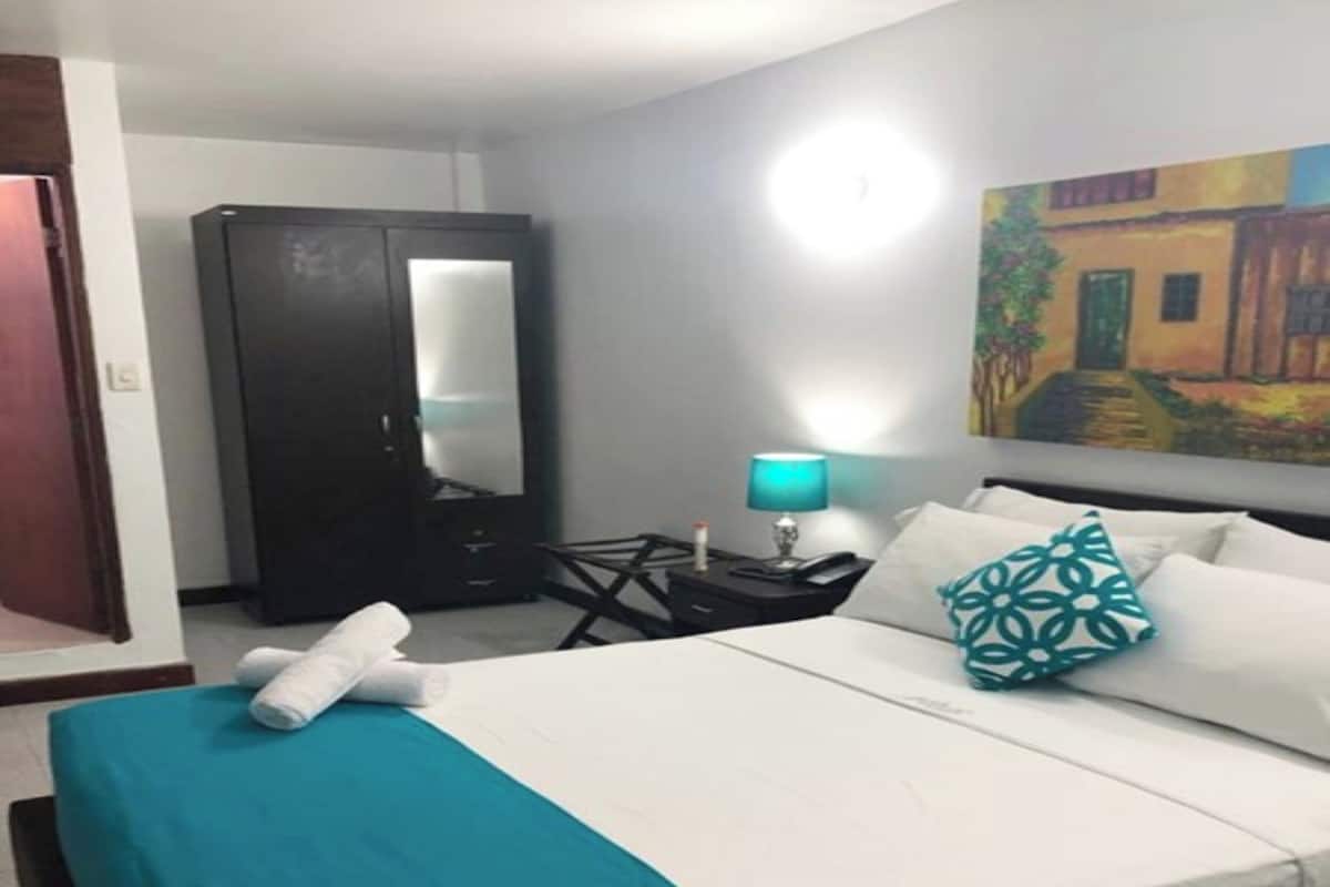 A hotel room with a bed, a desk, a chair, a wardrobe, and a painting on the wall.