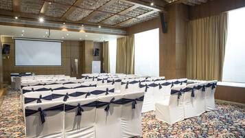 Un salón de conferencias con filas de sillas blancas atadas con cintas azules, una pantalla de proyección grande y suelo alfombrado.