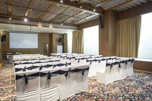 Un salón de conferencias con filas de sillas blancas atadas con cintas azules, una pantalla de proyección grande y suelo alfombrado.