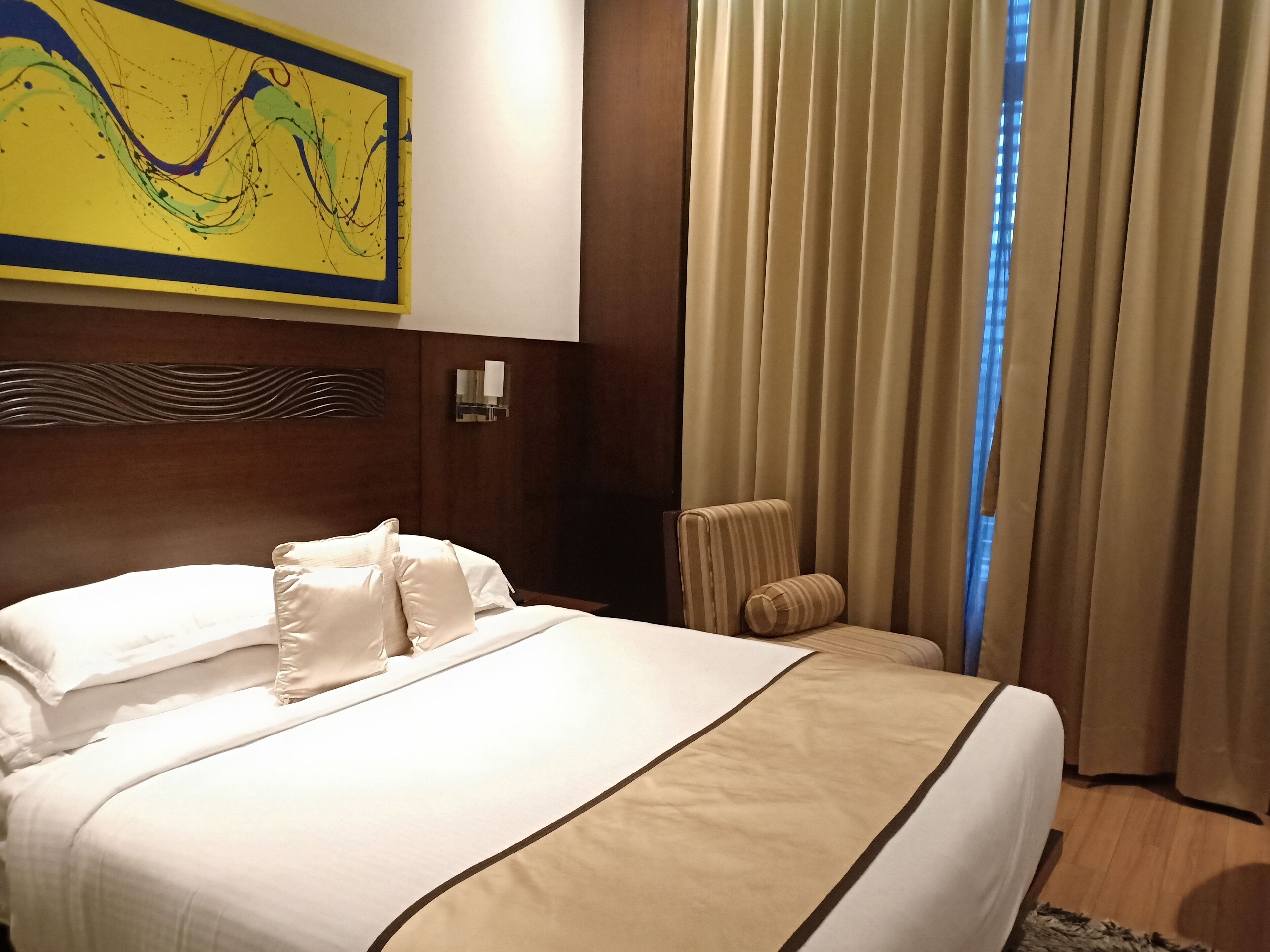 A hotel room with a large bed, a headboard with a wavy design, a wall-mounted light fixture, and a window with curtains.