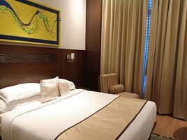 A hotel room with a large bed, a headboard with a wavy design, a wall-mounted light fixture, and a window with curtains.