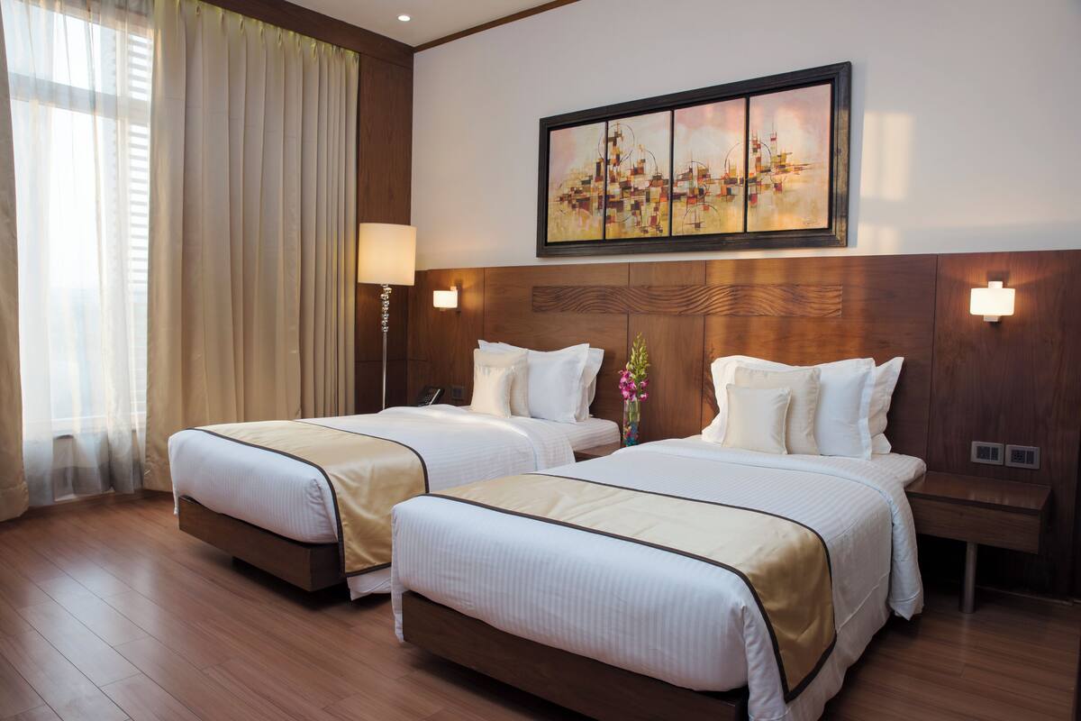 Habitación de hotel con dos camas, un cuadro grande en la pared, cabecera de madera y mesita de noche con lámpara.