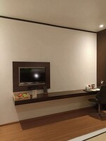 Une chambre d’hôtel moderne dotée d’un téléviseur à écran plat fixé sur un panneau en bois foncé, d’un petit bureau avec une chaise et d’un plancher en bois.