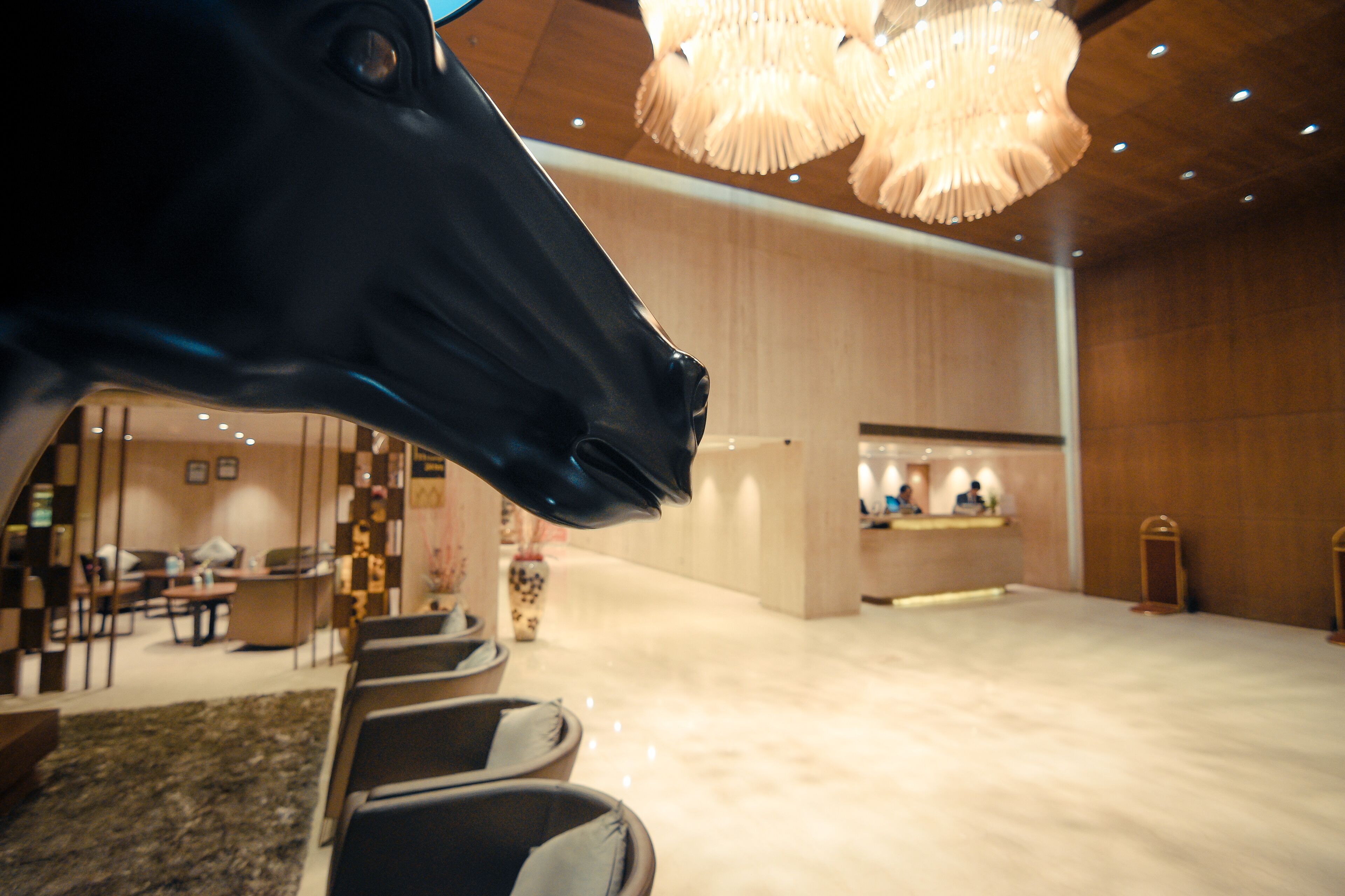 A modern hotel lobby with a large black horse sculpture, a reception desk, and a seating area with chairs and tables.