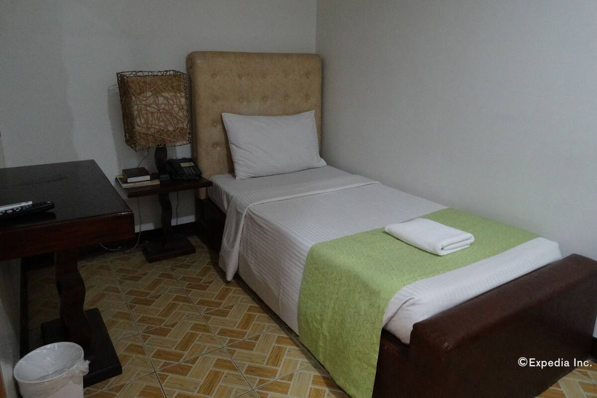 A small, neatly arranged hotel room with a single bed, a bedside table with a lamp, a small desk with a remote control, and a trash bin.