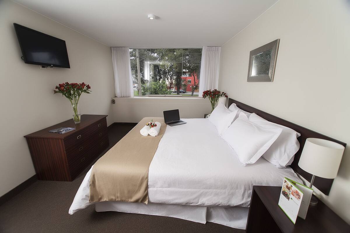 Una habitación de hotel con cama, escritorio, laptop, televisor y ventana con cortinas.