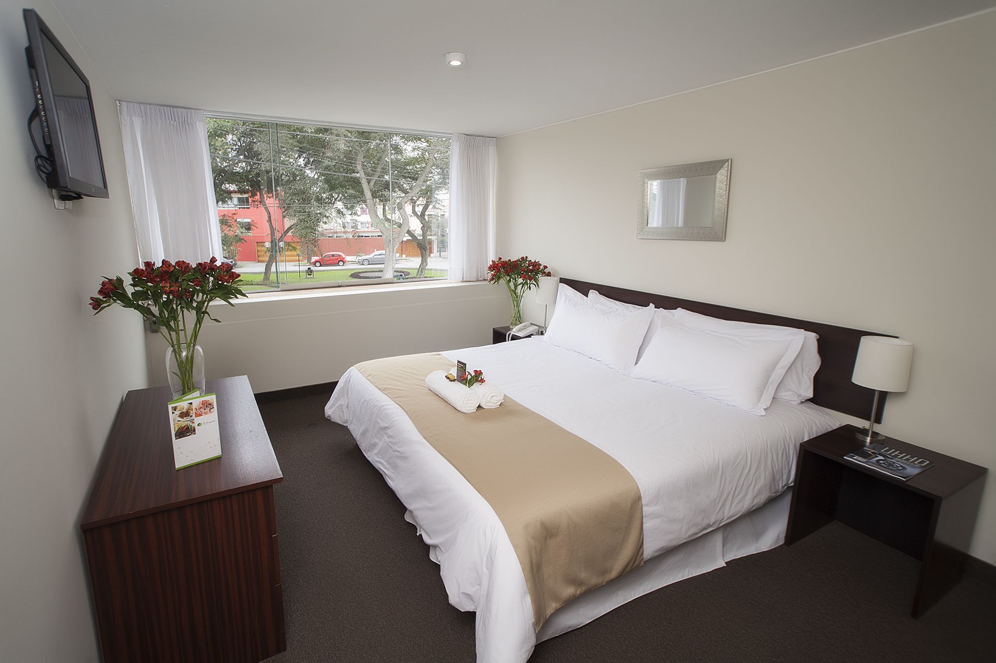 Una habitación de hotel con cama, mesita de noche, televisor, ventana con cortinas y un jarrón con flores.